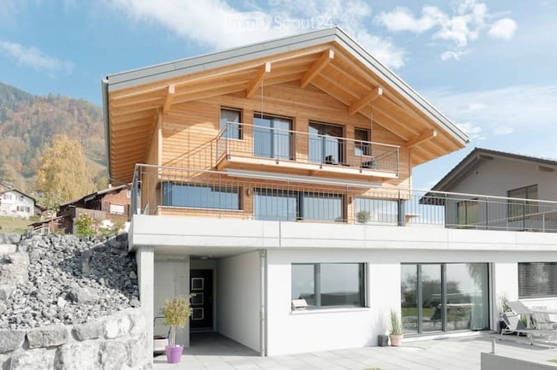 Modern two-story house with wooden facade, large windows, and a balcony overlooking a scenic mountain view.