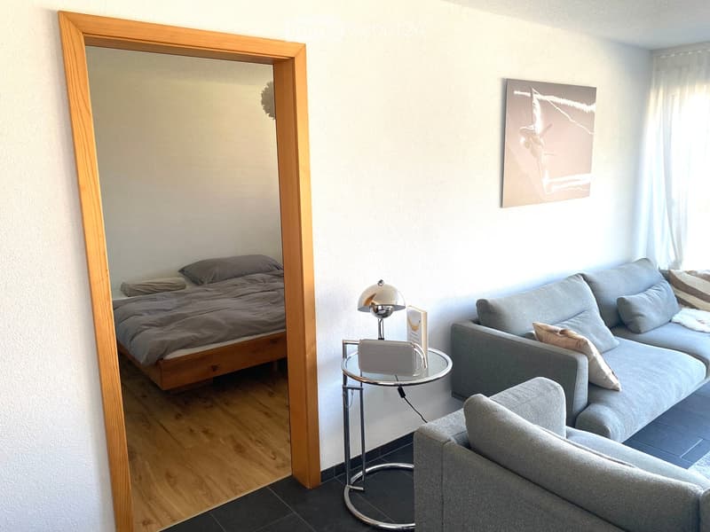 A modern, well-lit room with a grey sofa, a wooden bed, a round glass table with a lamp, and a large mirror. The room features a contemporary aesthetic with neutral tones and a mix of wooden and metallic finishes.