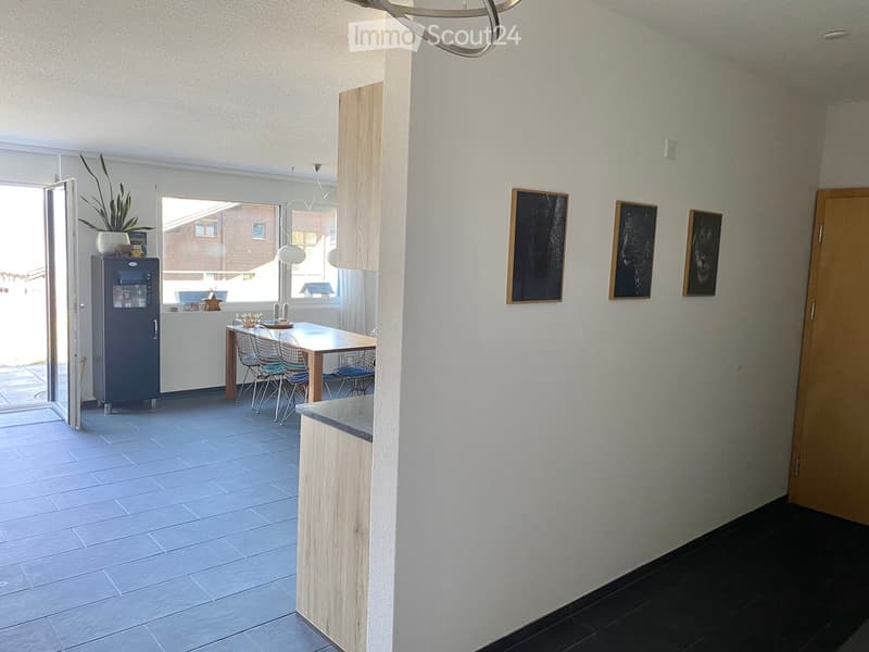 Modern kitchen and dining area with light-colored tiles, a wooden dining table, four chairs, a refrigerator, and framed pictures on the wall.