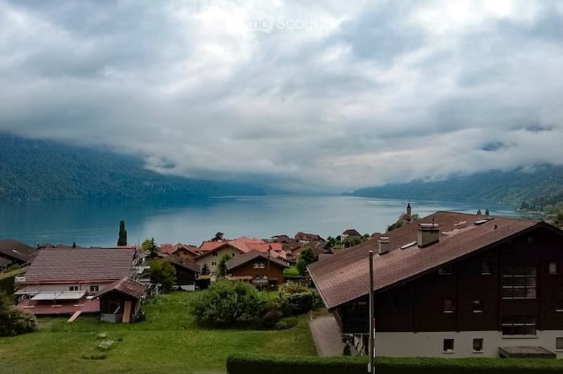 A scenic view of a small village nestled by a lake, surrounded by mountains under a cloudy sky. Traditional wooden houses with pitched roofs are scattered across the landscape.