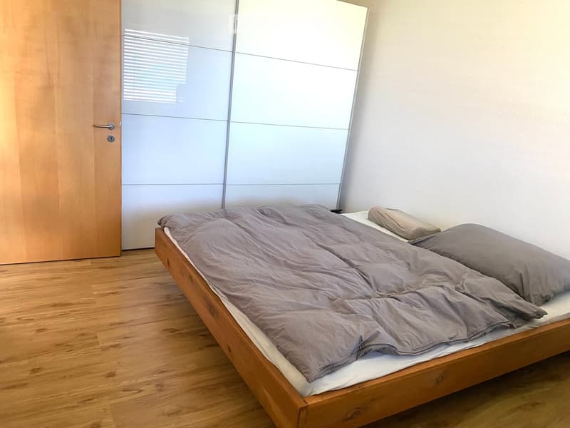 Minimalist bedroom with a wooden bed frame, grey duvet, and two pillows, featuring a sliding wardrobe and wooden flooring.