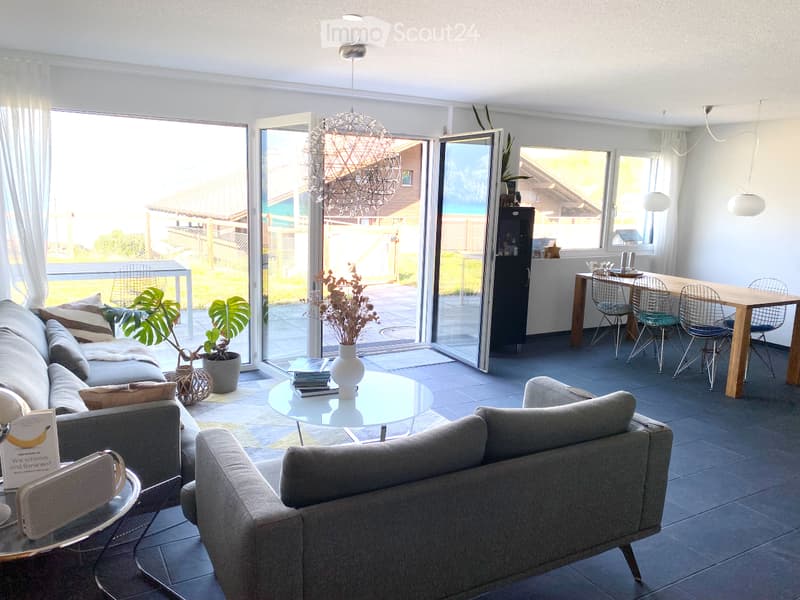 Modern living and dining area with large windows and sliding glass doors leading to a patio. Features a grey sectional sofa, glass coffee table, dining table with chairs, and decorative plants.