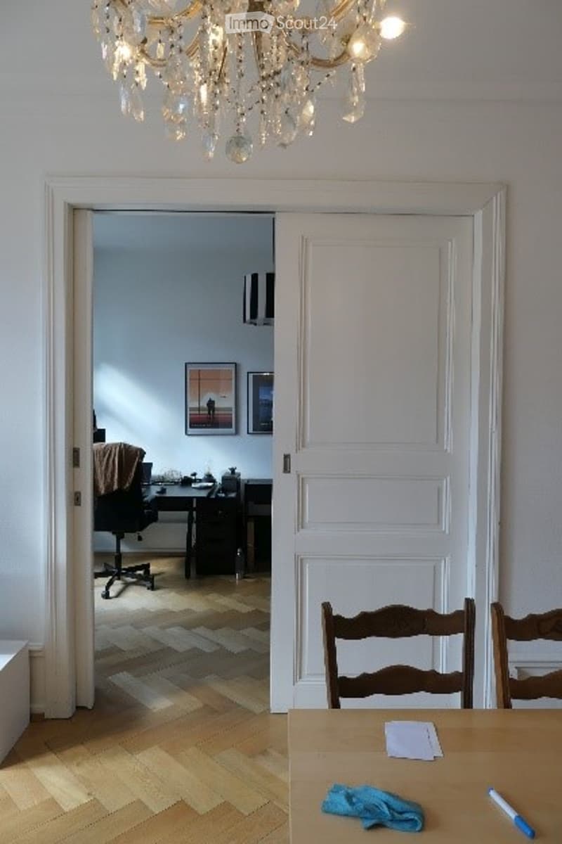 A well-lit room with wooden flooring, a chandelier, a table with chairs, and an open doorway leading to a study area with a desk, chair, and laptop.