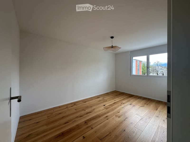 Empty room with wooden flooring, white walls, and a single window with a view of the outside. A hanging light fixture is visible on the ceiling.
