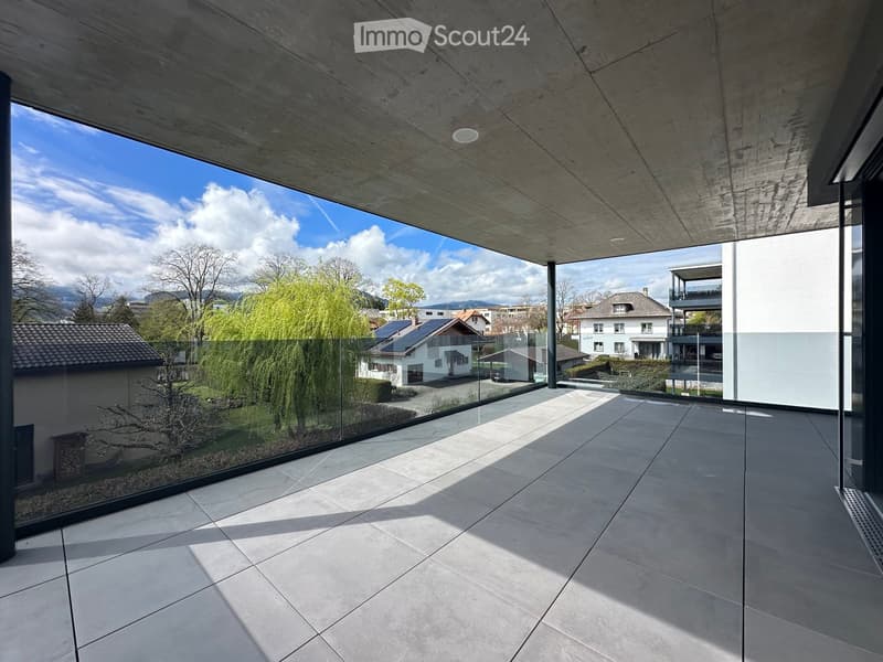 Modern balcony with large glass walls, concrete ceiling, and tiled floor, offering a view of a suburban neighborhood with trees and houses.