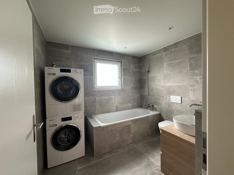 Modern bathroom with a bathtub, sink, toilet, and a stacked washing machine. Features grey tiled walls and a window for natural light.