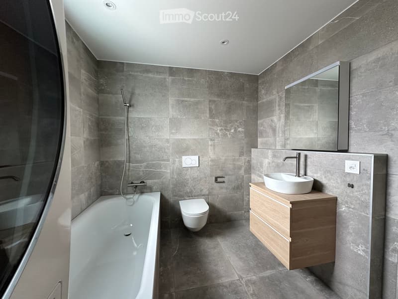 Modern bathroom with grey tiled walls and floor, featuring a bathtub, toilet, sink with a wooden vanity, and a large mirror above the sink.