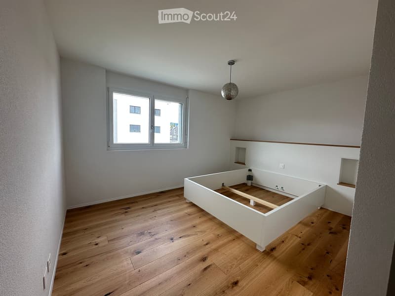 Empty room with wooden flooring, white walls, and a large window. A raised platform is present in the center, likely for a bed frame.
