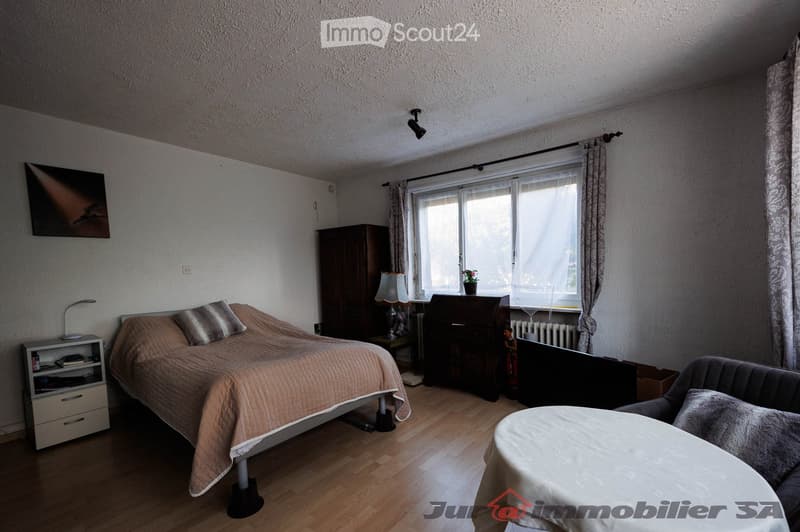 A well-lit bedroom with a bed, nightstand, lamp, sofa, table, cabinet, and a window with curtains. The room features wooden flooring and a radiator under the window.