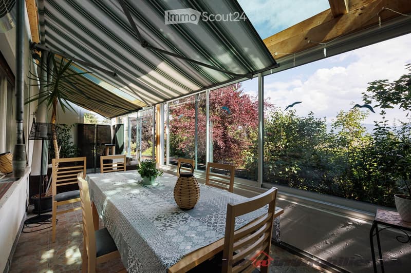 Brightly lit conservatory with large glass windows, wooden dining table with chairs, wicker basket centerpiece, floor lamp, and potted plants.