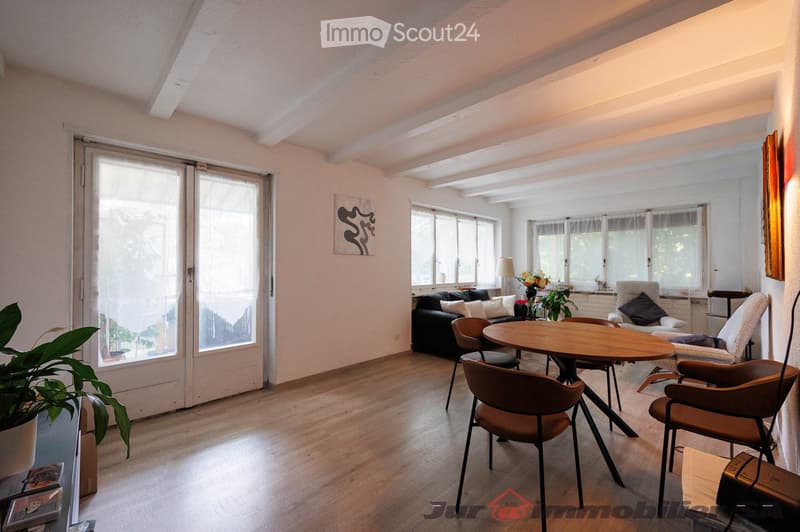Bright living room with wooden flooring, large windows, and sliding glass doors leading to a balcony. Features a black sofa, wooden dining table with chairs, a floor lamp, and decorative plants.