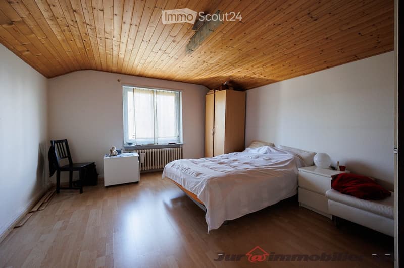 Cozy bedroom with wooden flooring, slanted wooden ceiling, large window, bed with white bedding, nightstand, wardrobe, and a small table with a chair.
