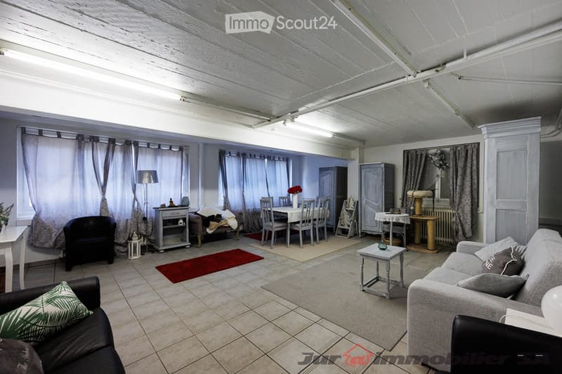 A spacious, well-lit living and dining area with exposed wooden beams on the ceiling. The living area features a grey sofa, black armchair, coffee table, and a small side table. The dining area includes a white dining table with six chairs, a sideboard, a