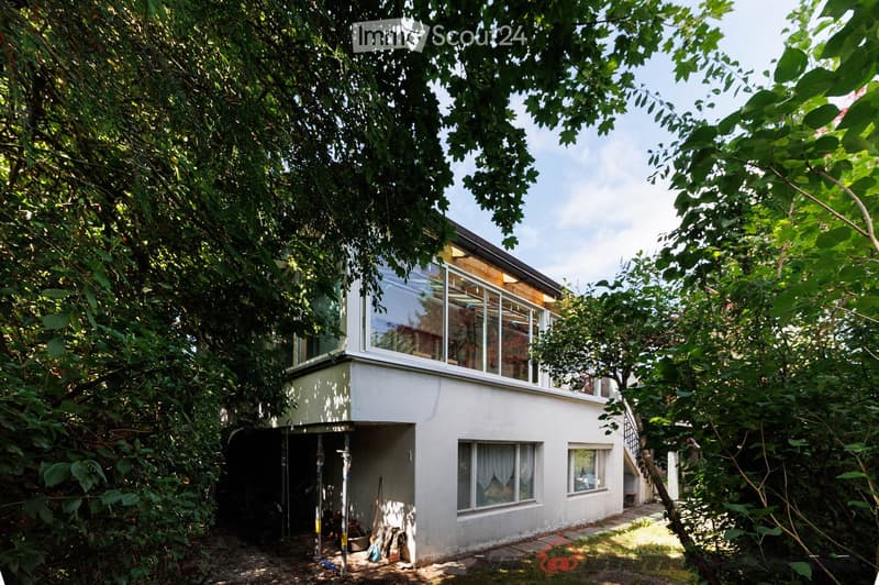 A two-story modern house with white walls, large windows, and a raised ground floor, surrounded by lush greenery and trees.
