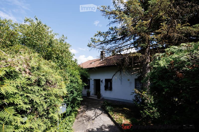 A small, single-story white house with a tiled roof, surrounded by lush greenery and trees. The house features a front door with a small porch and windows, and is accessed via a stone pathway.