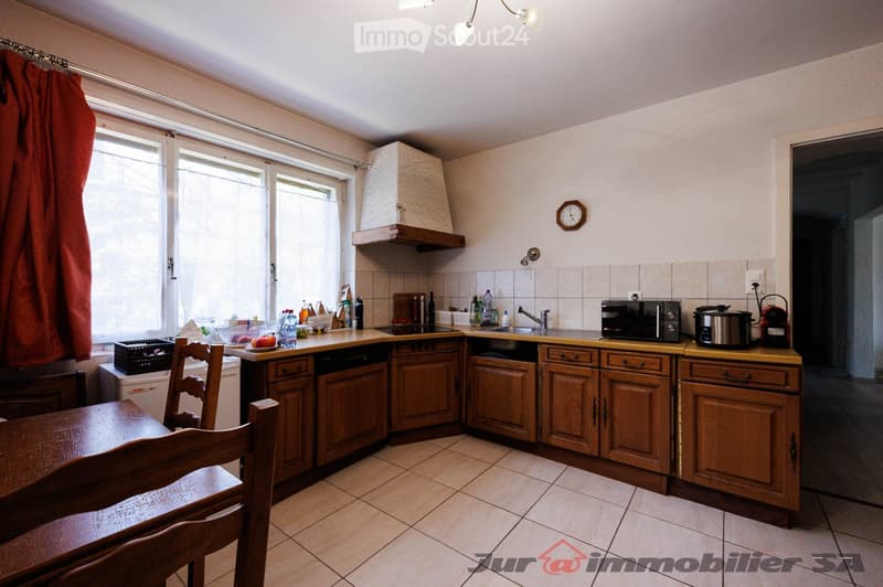 A well-lit kitchen with wooden cabinets, a sink, microwave, pot, and a small dining area with a table and chairs. The kitchen features a window with a curtain and a clock on the wall.