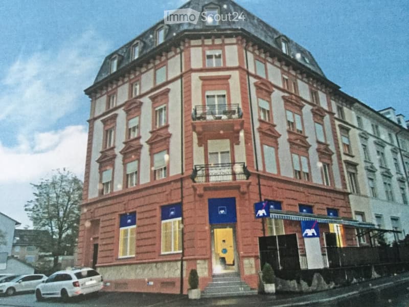 A multi-story building with red brick facade, multiple windows, balconies, and a central entrance with a yellow door.
