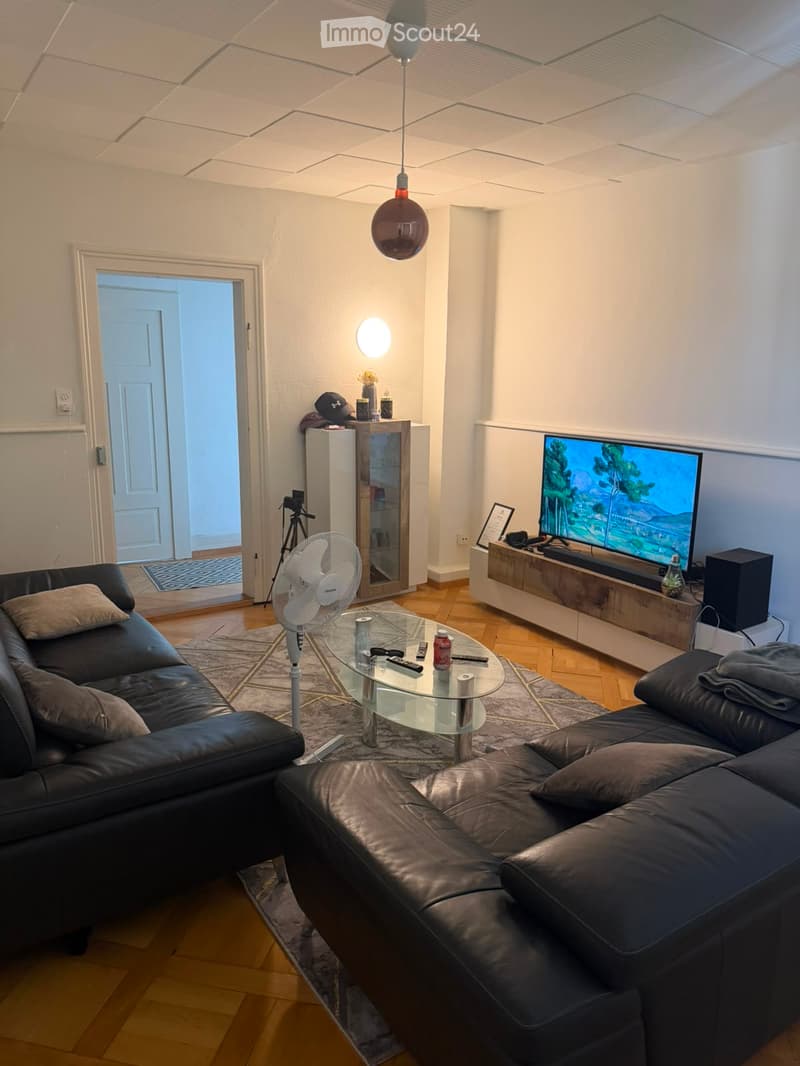 Modern living room with a black leather sofa, glass coffee table, TV, fan, and a small cabinet with decorative items.
