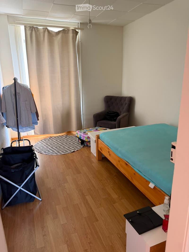 Cozy bedroom with wooden flooring, a single bed with blue bedding, a small wooden chair, a round patterned rug, a clothing rack with shirts, a small table with a laptop and bottle, and a colorful stool near the window.