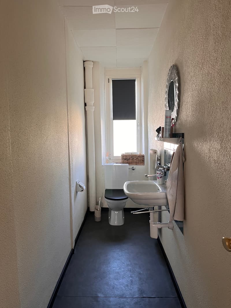 Modern bathroom with a toilet, sink, mirror, and towel. Features a window with blinds and a small shelf with personal care items.