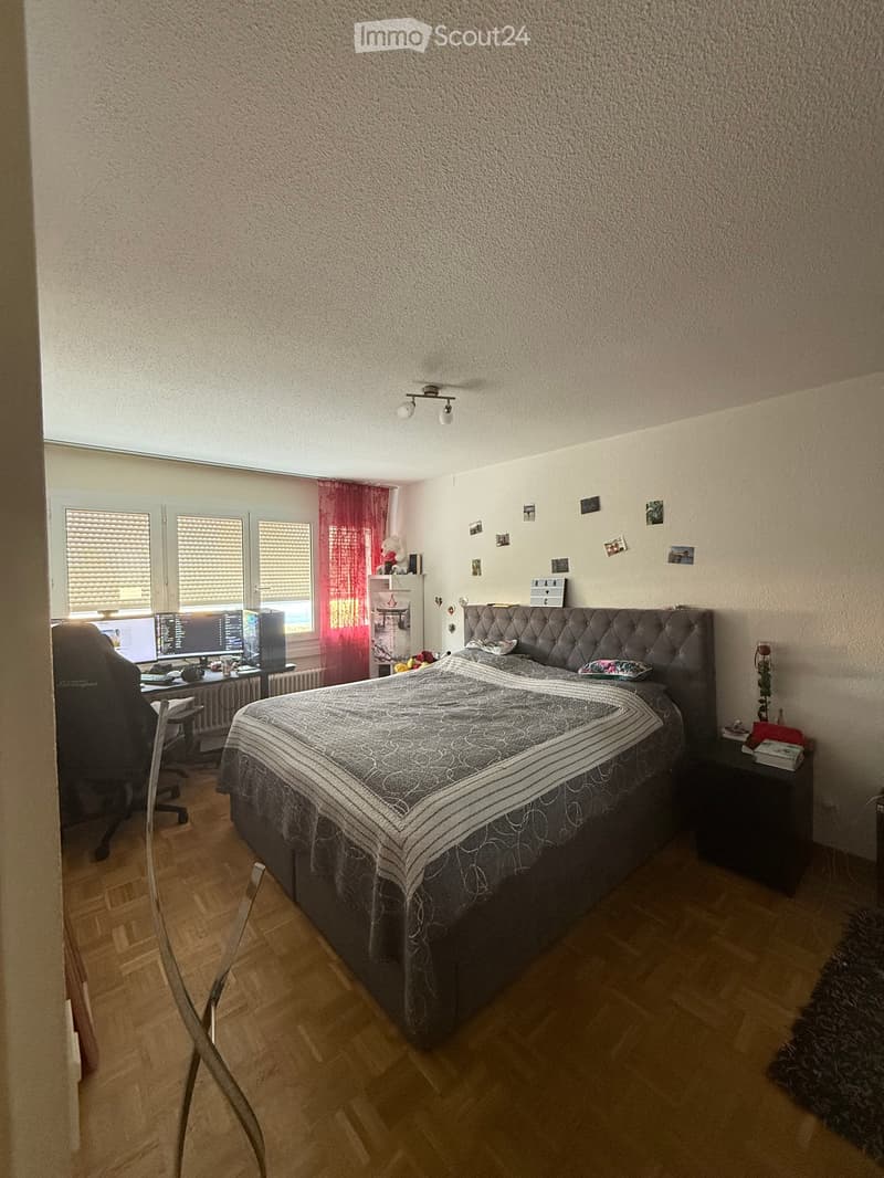 A well-lit bedroom with a large bed, nightstand, desk with computer, and a window with red curtains.