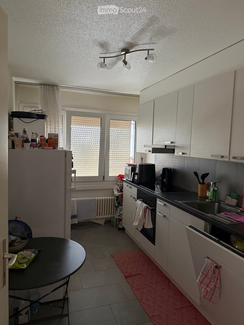 Modern kitchen with white cabinets, black countertops, and stainless steel appliances. Features include a refrigerator, microwave, coffee maker, oven, sink, and a small round table with a chair. The space is well-lit with natural light from a window with