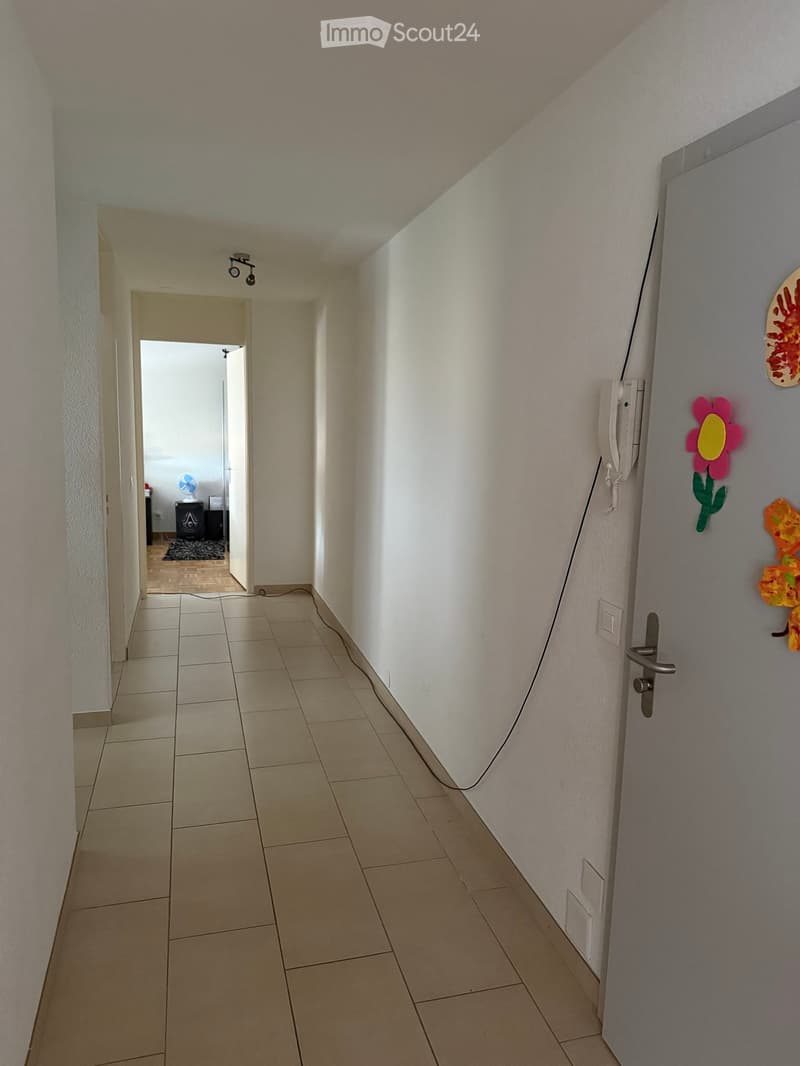 A clean, tiled hallway with white walls, a closed door on the right, and an open doorway leading to another room on the left. The hallway has a light fixture on the ceiling and a small fan visible in the adjacent room.