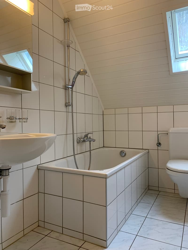 Modern bathroom with white tiles, a bathtub, toilet, sink, and showerhead. Features include a sloped ceiling and built-in shelves.