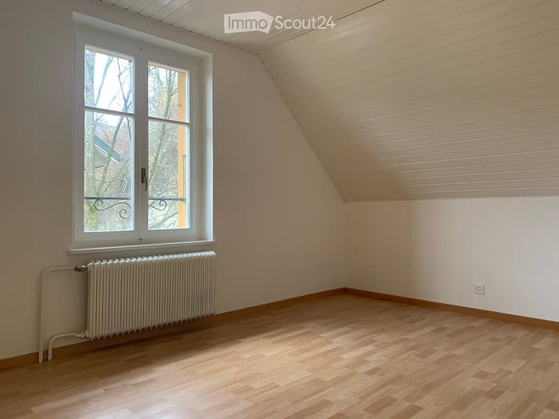 Empty attic room with wooden flooring, white walls, and a large window with a radiator below it.
