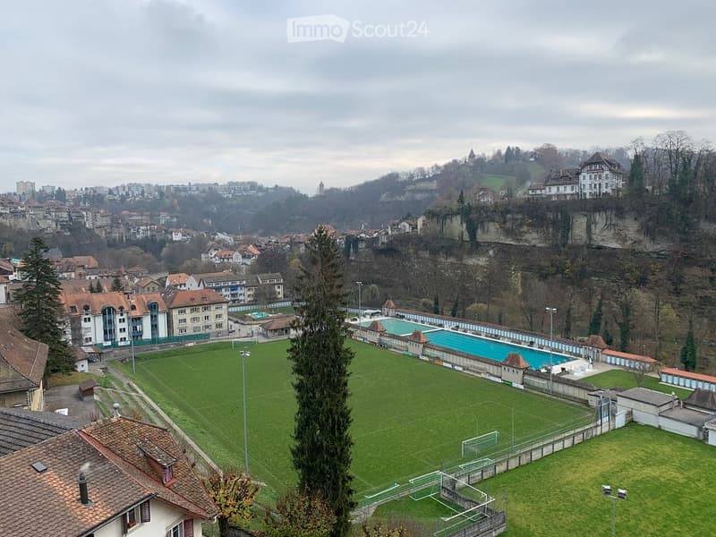 A panoramic view of a cityscape with a large green sports field, a swimming pool, and surrounding buildings.