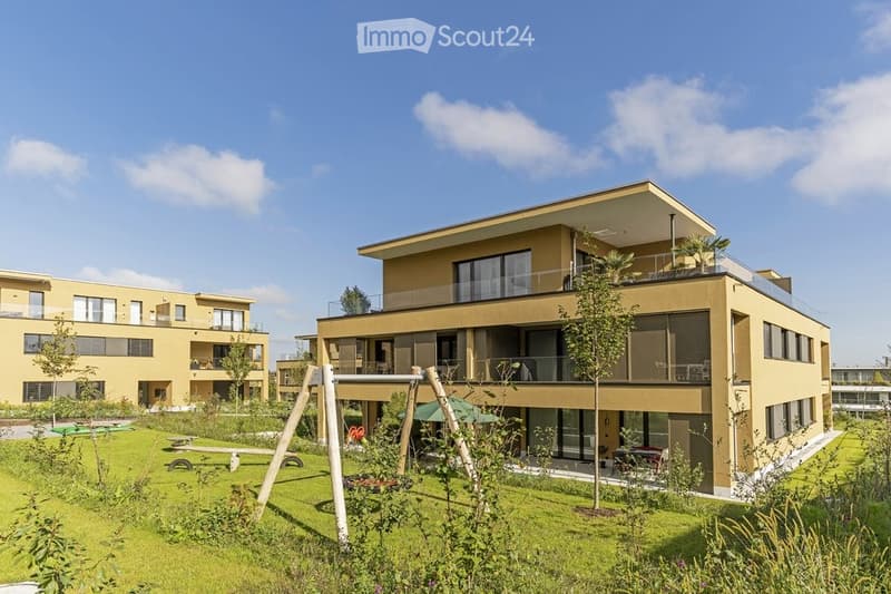 Modern two-story residential building with large windows, balconies, and a landscaped garden area with playground equipment.