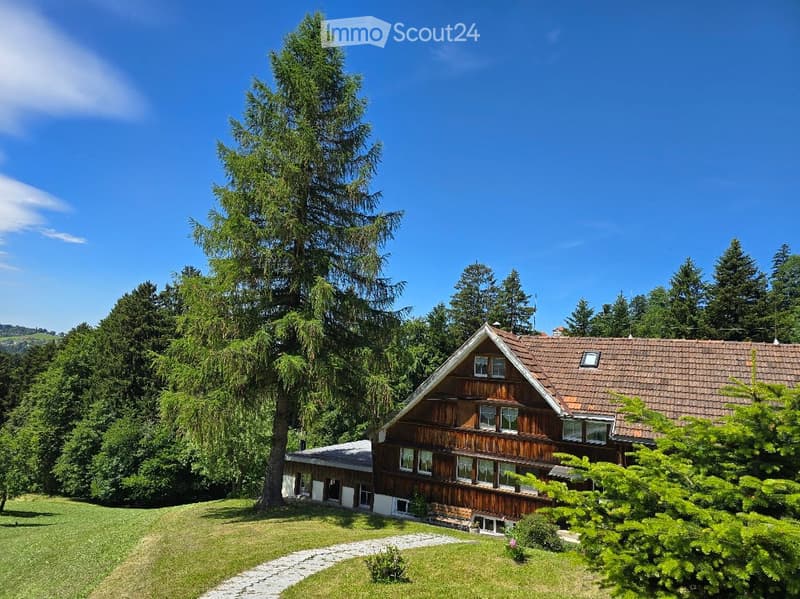 A two-story wooden house with a red-tiled roof, surrounded by lush greenery and trees, with a stone pathway leading to the entrance.