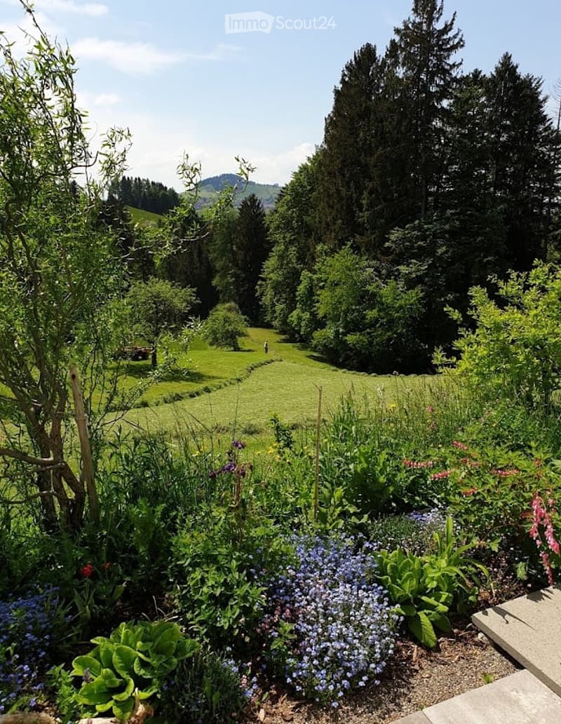 A lush garden with a variety of flowers, shrubs, and trees, set against a backdrop of green hills and tall coniferous trees. The garden features a well-maintained lawn and a winding path.