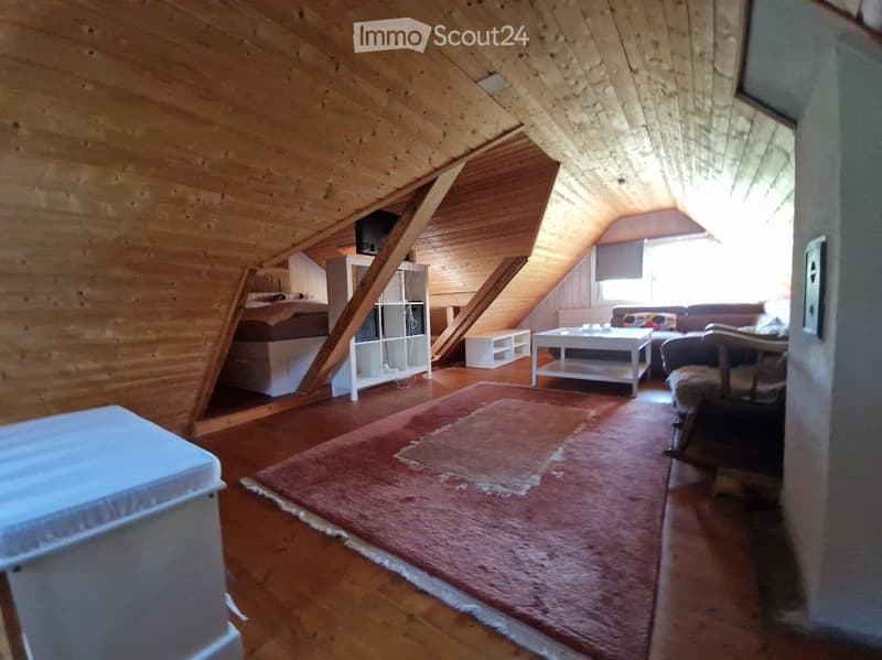 Cozy attic room with wooden walls and ceiling, featuring a sofa, coffee table, chair, shelf, and bed.