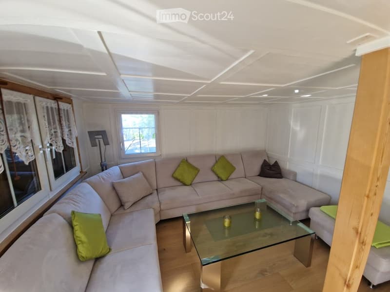 Modern living room with a grey sectional sofa, glass coffee table, ottoman, and floor lamp. The room features white paneling and large windows with curtains.