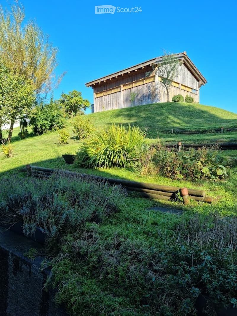 A wooden house sits atop a grassy hill, surrounded by lush greenery and plants. The scene includes a variety of shrubs, trees, and a wooden fence along the pathway.