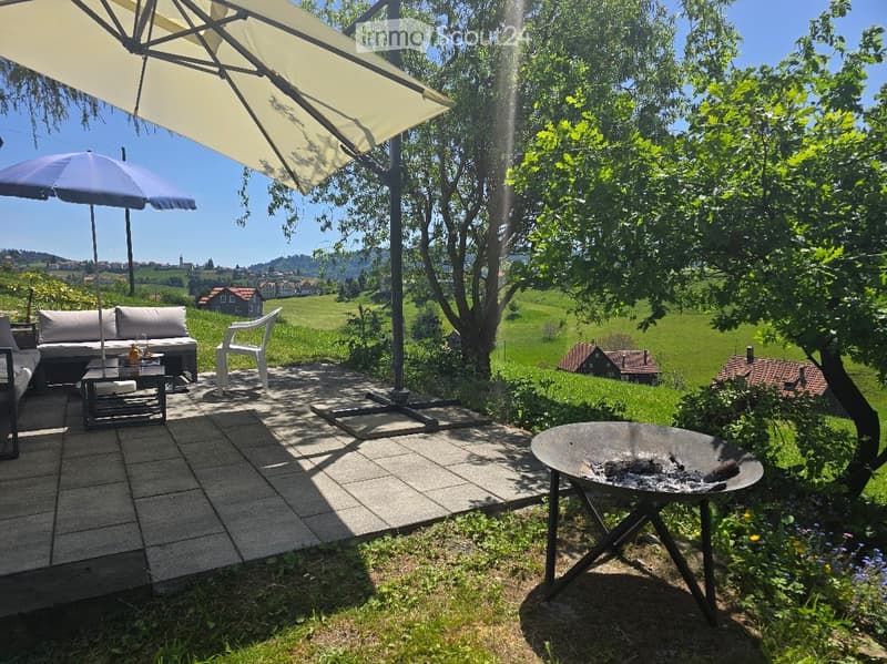 Outdoor patio with a sofa, coffee table, chairs, large umbrella, and a fire pit, overlooking a scenic countryside view.