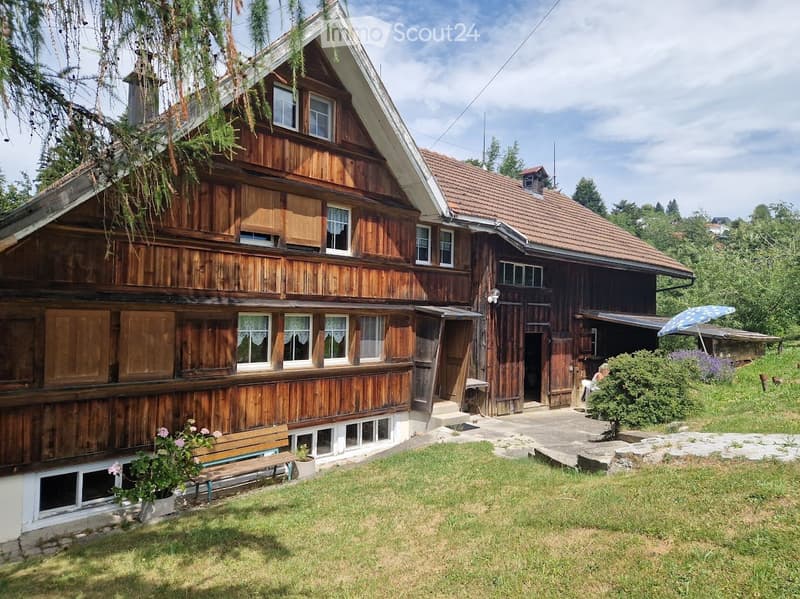 Traditional wooden house with a red-tiled roof, multiple windows, and a small garden area with a bench and potted plants.