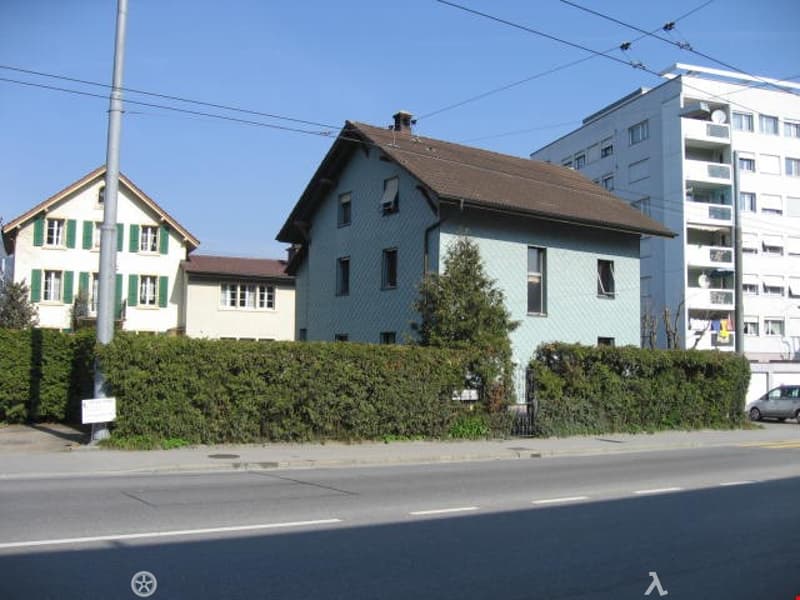 A two-story residential house with a light blue exterior and brown roof, surrounded by greenery and situated near a road with other buildings in the background.