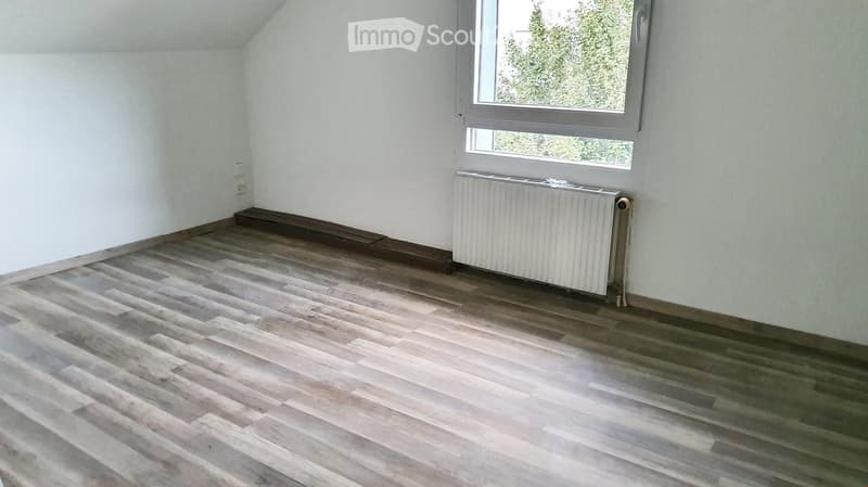 Empty room with wooden flooring, white walls, and a radiator. A window with a view of greenery is visible.