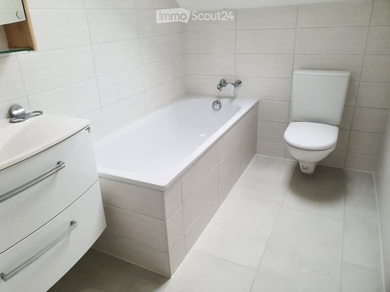 Modern bathroom with white tiles, a bathtub, a toilet, a sink with a cabinet, and a glass shelf above the sink.