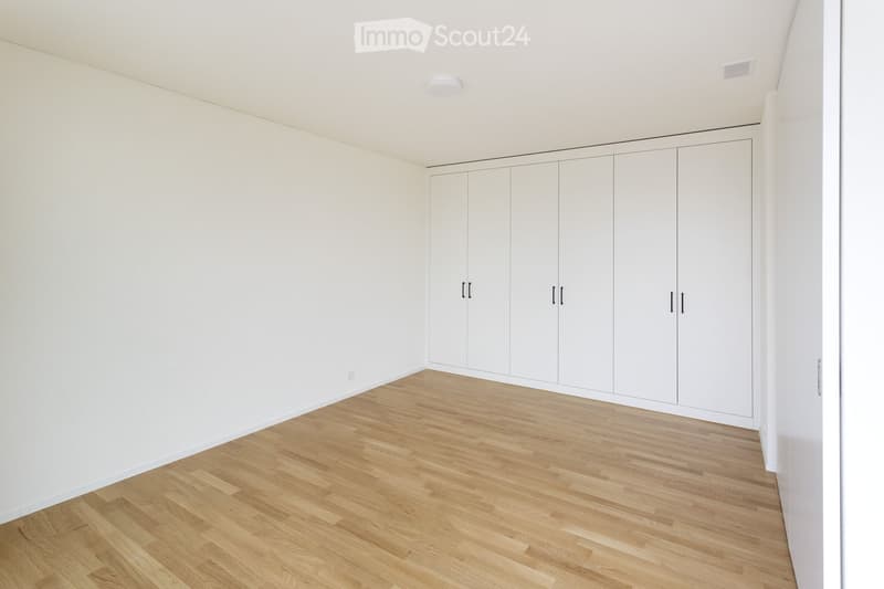Empty room with white walls, light wooden flooring, and built-in white wardrobes along one wall.