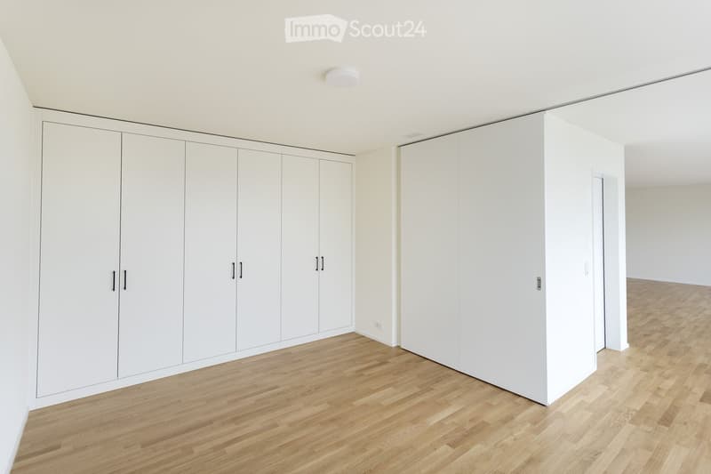 Empty room with light wooden flooring, white walls, and built-in white wardrobes with black handles.