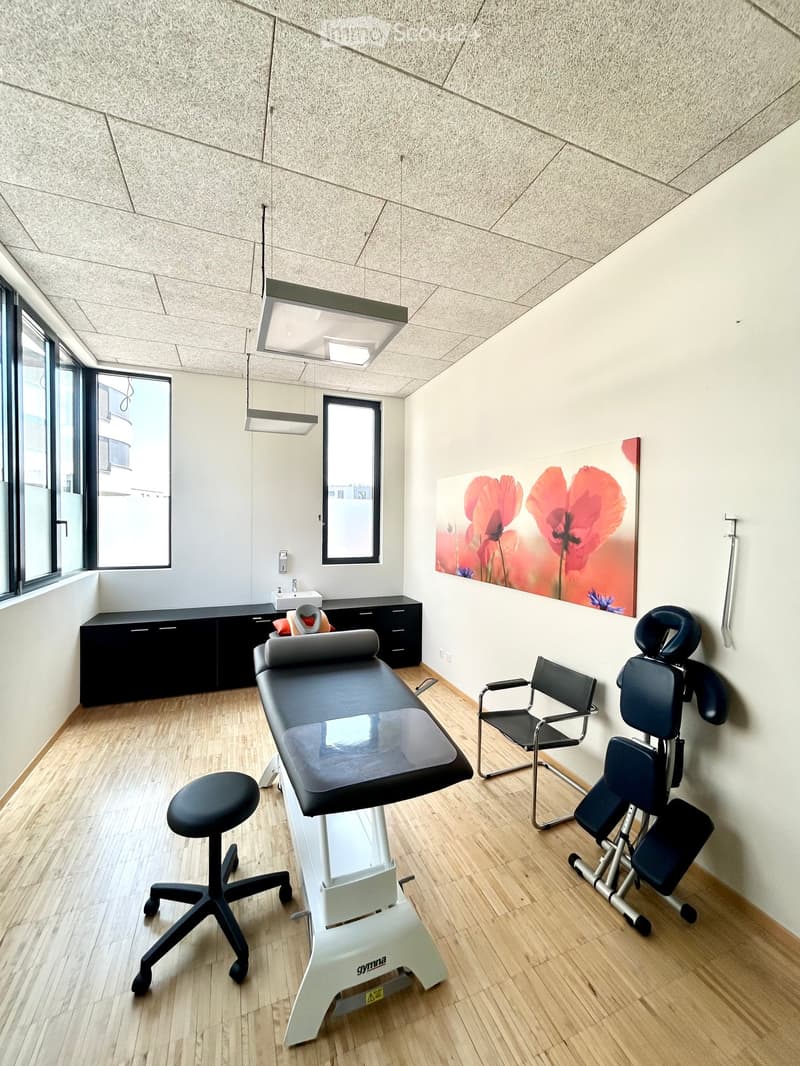 Modern medical examination room with wooden flooring, exam table, stool, chairs, cabinet, sink, and artwork on the wall.