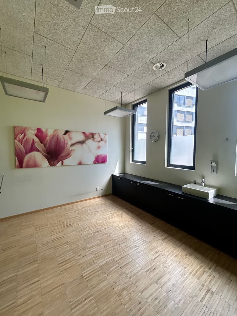 Empty room with wooden flooring, modern lighting fixtures, and a sink counter with a faucet. The walls are light-colored with a large floral painting and a clock.