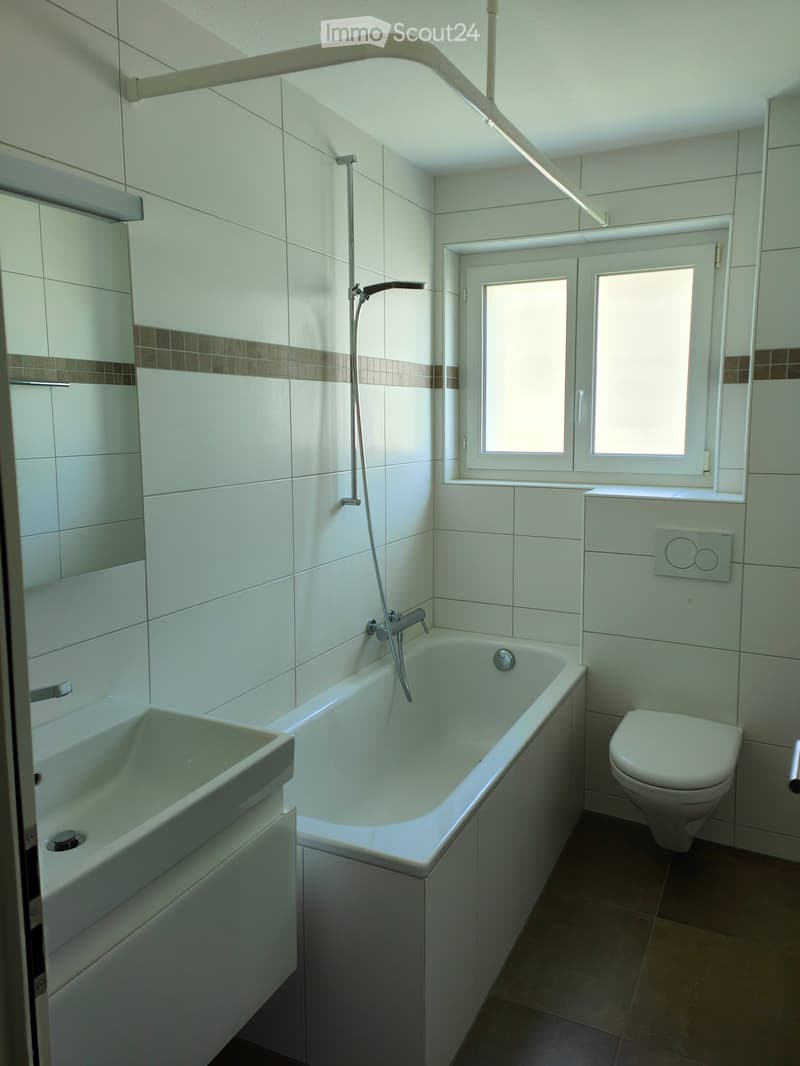 Modern bathroom with white tiles, a bathtub, a toilet, a sink, and a showerhead. The room features a window with frosted glass panes and a small cabinet next to the sink.