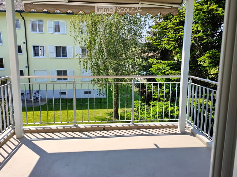 Balcony with metal railing, concrete floor, and a view of a green garden and a neighboring building with shutters.