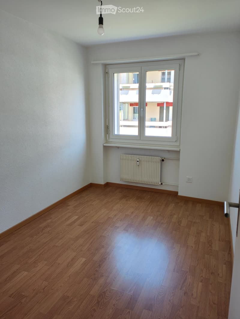 Empty room with wooden flooring, white walls, a window with white frame, and a radiator below the window.