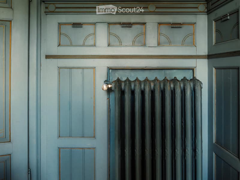 A hallway with light blue walls, ornate gold-framed doors, and a decorative radiator with a vintage design.