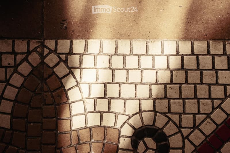 Close-up view of a patterned tile floor in a hallway, featuring a mix of beige, brown, and white tiles arranged in a geometric design.
